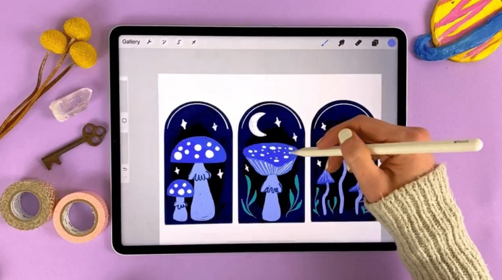 A person's hand drawing in the Procreate app on an iPad. The iPad is resting on a pink tabletop, and the drawing depicted is of three purple mushrooms under a moon and stars.