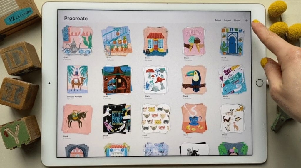 The Procreate app opened on an iPad. A person's hand is positioned over it, with her index finger hovering over a ‘plus’ sign icon in the upper right corner.