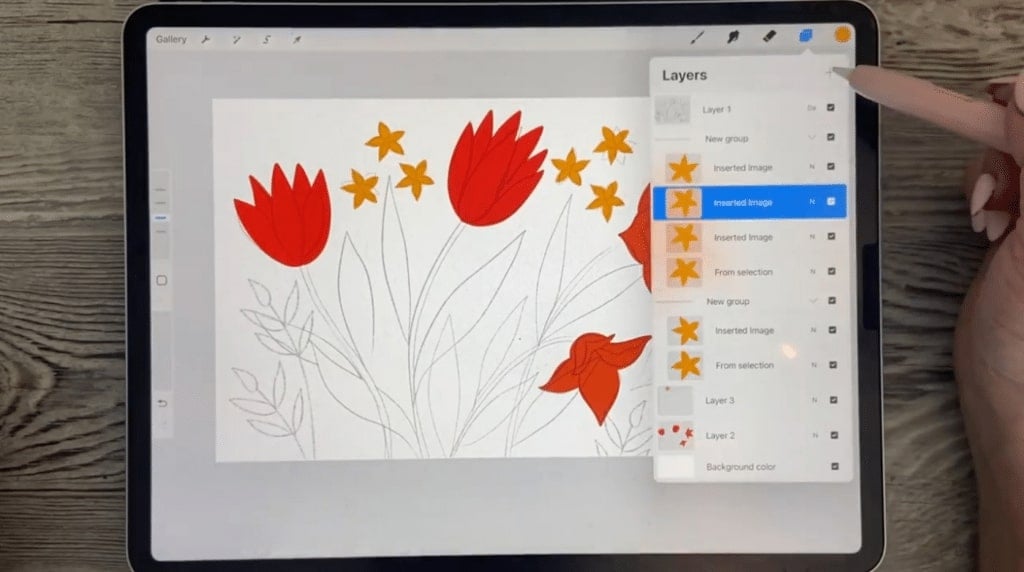 The Procreate app opened on an iPad, with a floral design on the canvas. A person's hand is using a stylus to open a menu titled ‘Layers’ in the upper right corner.