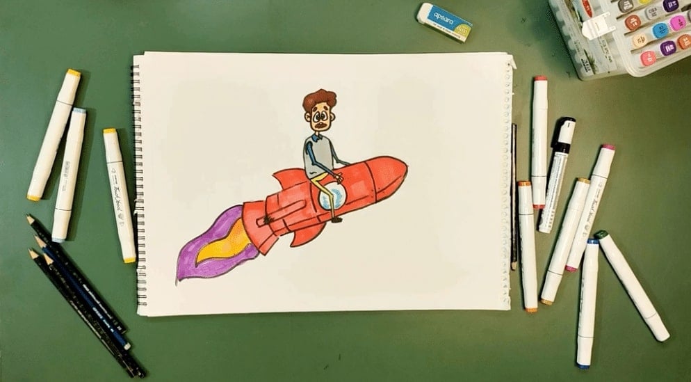 A male cartoon character riding a red rocket with purple and gold flames, with the sketchbook placed on a green table surrounded by colored pens.