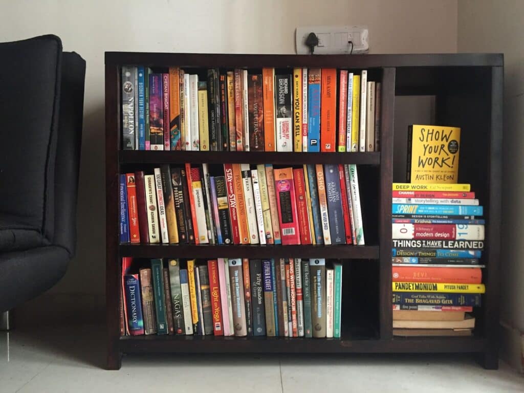 A black armchair sits next to a dark wooden shelving unit with three horizontal shelves to the left and one tall vertical shelf to the right. The shelves are completely full with a variety of different colored books. 