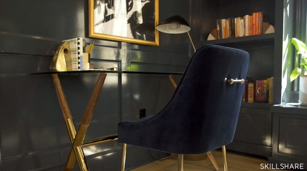 A blue velvet chair sits in front of a glass and gold desk. Next to it is a small built-in bookshelf filled with books and two bookends. 