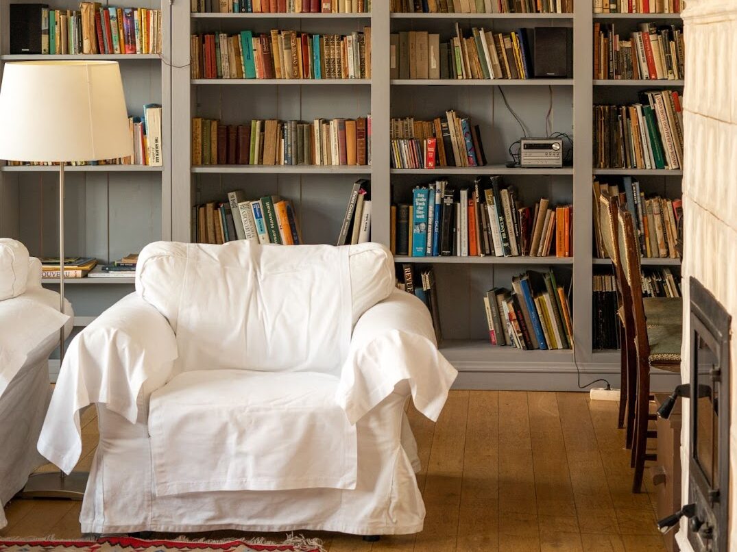 A small white couch sits behind a towering wall lined with gray bookshelves. Every shelf is filled with books besides one that contains a small, vintage silver stereo. A lamp, two chairs and a wood burning stove are also visible. 
