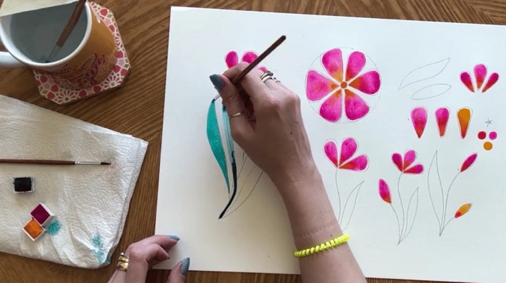 A person's hand painting bright pink and green watercolor flowers on a white piece of paper.