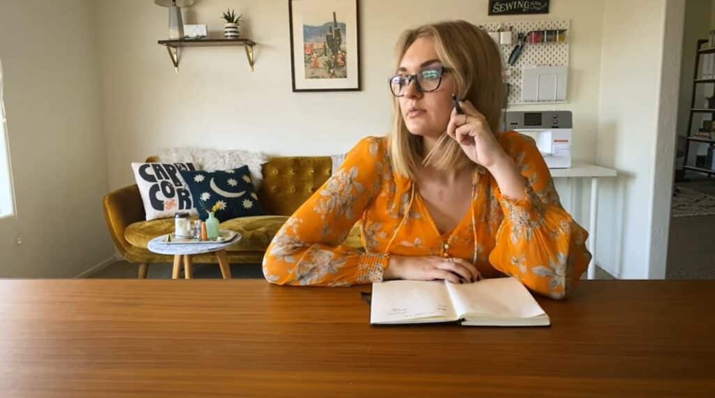 A woman wearing an orange dress and glasses sits at a wooden table with an open journal in front of her.