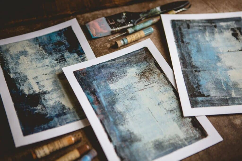 Three monoprints laying on a wooden surface. They each depict abstract images in shades of blue, white, black, and brown.