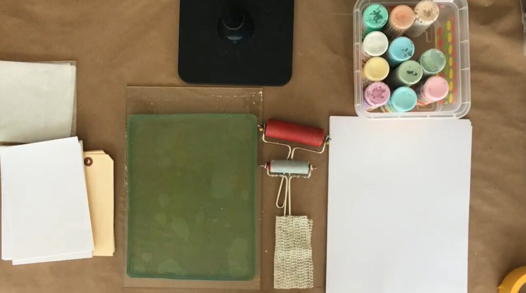 Various art supplies laid out on a brown paper surface, including two rollers, a gelli plate, paper and a plastic bin full of paints.