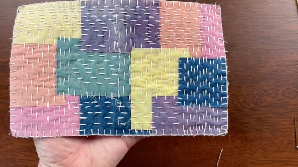 A hand holds a colorful patchwork rectangle covered in white, multidirectional stitches. 