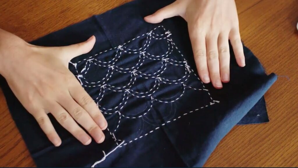Two hands press a piece of dark blue fabric onto a wooden table. On the fabric is a scalloped design stitched in white thread.