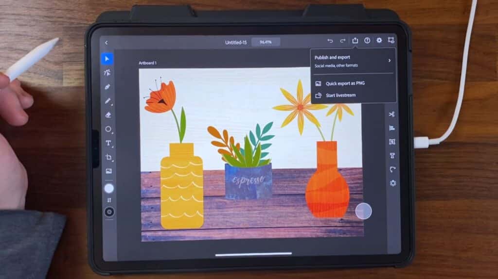 A person’s hand resting on a wood table holding an Apple Pencil. In the center of the image is an Apple iPad, which is displaying a vector image within Adobe Illustrator. The image depicts three vases, each filled with a plant or flower.
