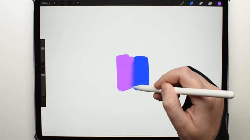 A person’s hand holding an Apple Pencil over an iPad lying on a white tabletop. The Procreate app is open on the iPad, and the person is using the Apple Pencil to blend a blue rectangle with a pink rectangle.