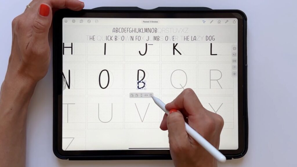 A person’s hand holding an Apple Pencil over an iPad on a white tabletop. They’re using the Apple Pencil to draw the letter B in a graphic design application Fontself. 