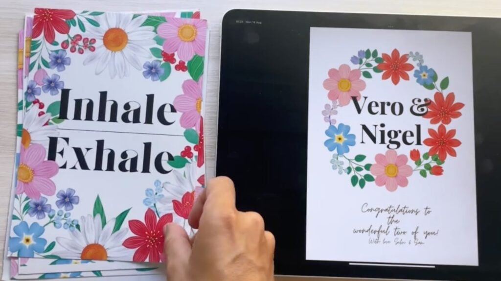 Printed posters and a tablet sit on a table. The posters have a floral frame and the words “Inhale, Exhale” on them. The tablet has a design with a circle of flowers surrounding the names “Vero & Nigel.”