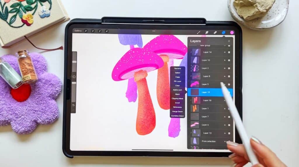 A person’s hand holding an Apple Pencil over an iPad with Procreate open on the screen. The Procreate canvas shows an illustration of bright neon pink and orange mushrooms, while the person uses their Apple Pencil to open the options for a specific layer. 