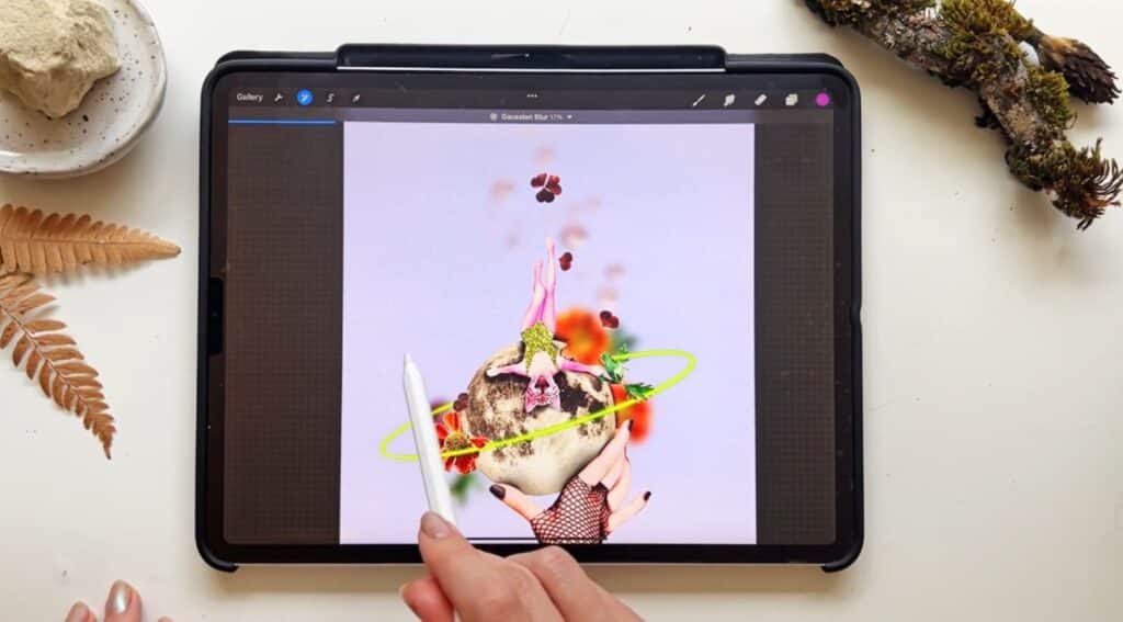 A person’s hand holding an Apple Pencil over an iPad on a white tabletop. The Procreate app is open on the iPad, and is displaying a colorful and surreal image of a cat, a moon and flowers on a lilac background.