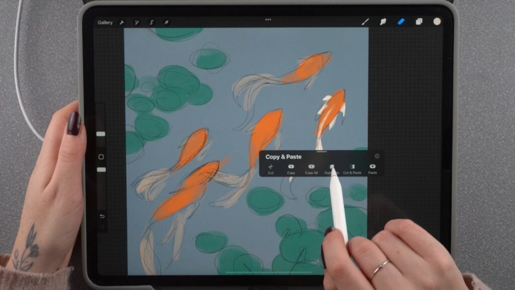 A person’s hand holding an Apple Pencil over an iPad with Procreate open on the screen. Procreate’s canvas shows an illustration of koi fish swimming in a pond, and the person is using their Apple Pencil to interact with a menu titled ‘Copy & Paste.’