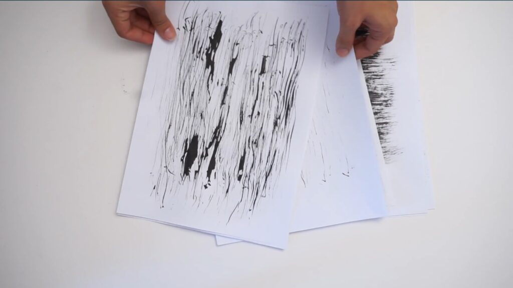 A person’s hands fanning out sheets of white paper on a white table. Each sheet of paper is decorated with a different minimalistic texture in black ink.