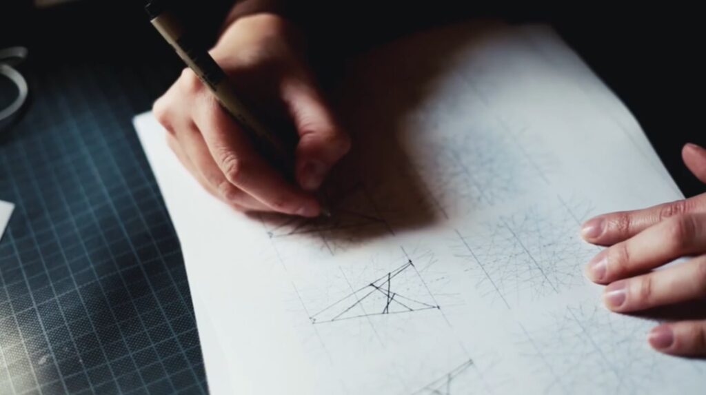 A person’s hand holding a pen and drawing on white paper on top of a dark-colored table. They’re using the pen to draw the letter A, which they’ll then use to form the first letter of their own typeface. 