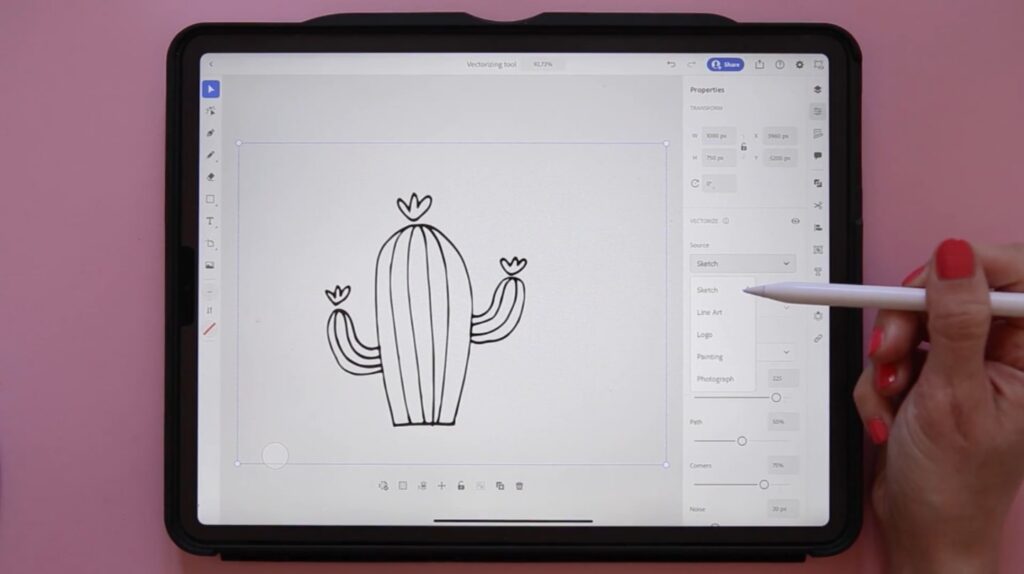 A person’s hand holding an Apple Pencil and resting on a pink tabletop. They’re using the pencil to tap on the screen of an iPad, which is displaying the program Adobe Illustrator which is being used to create a vector image of a saguaro cactus.