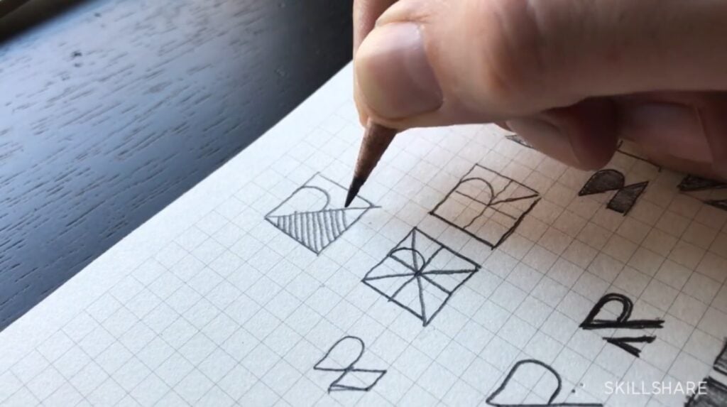 A sheet of white graphing paper on a black wooden tabletop. A person’s hand can be seen using a pencil to sketch several logos based on the letter ‘R.’