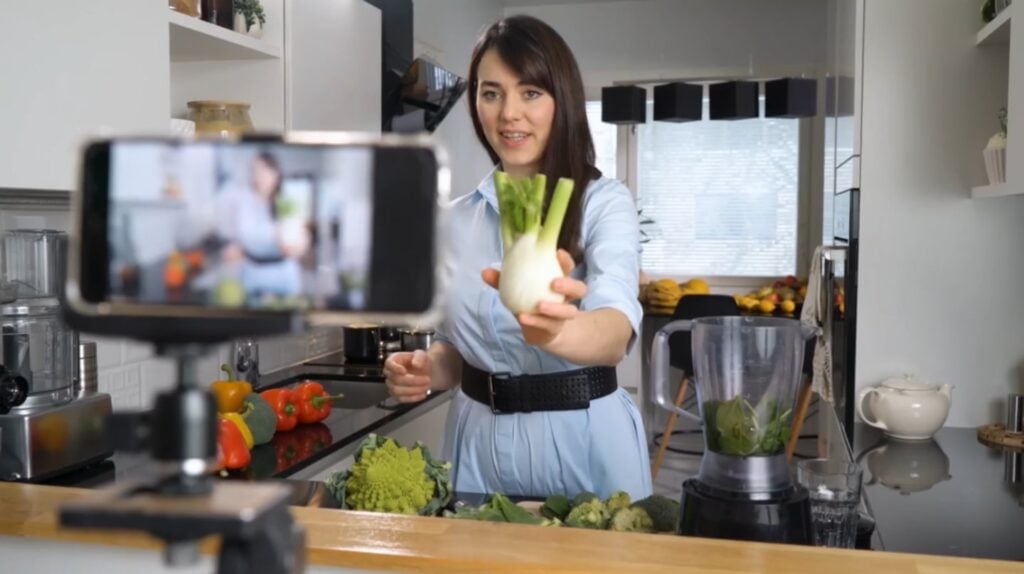 A woman in a blue dress standing in a kitchen. She is addressing a smartphone on a tripod that’s taking a video recording, and is holding a green and white vegetable up to the camera. 
