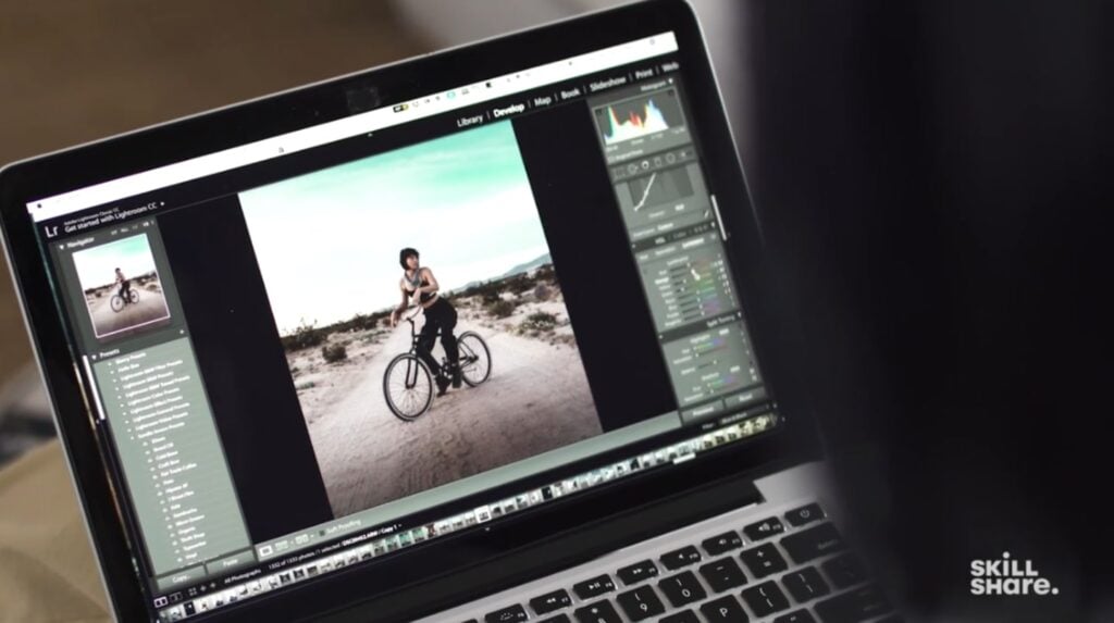 A MacBook laptop sitting open with the photo editing software Lightroom open on its screen. The software is being used to edit a photo of a person on a bike in the center of a long gravel road. 