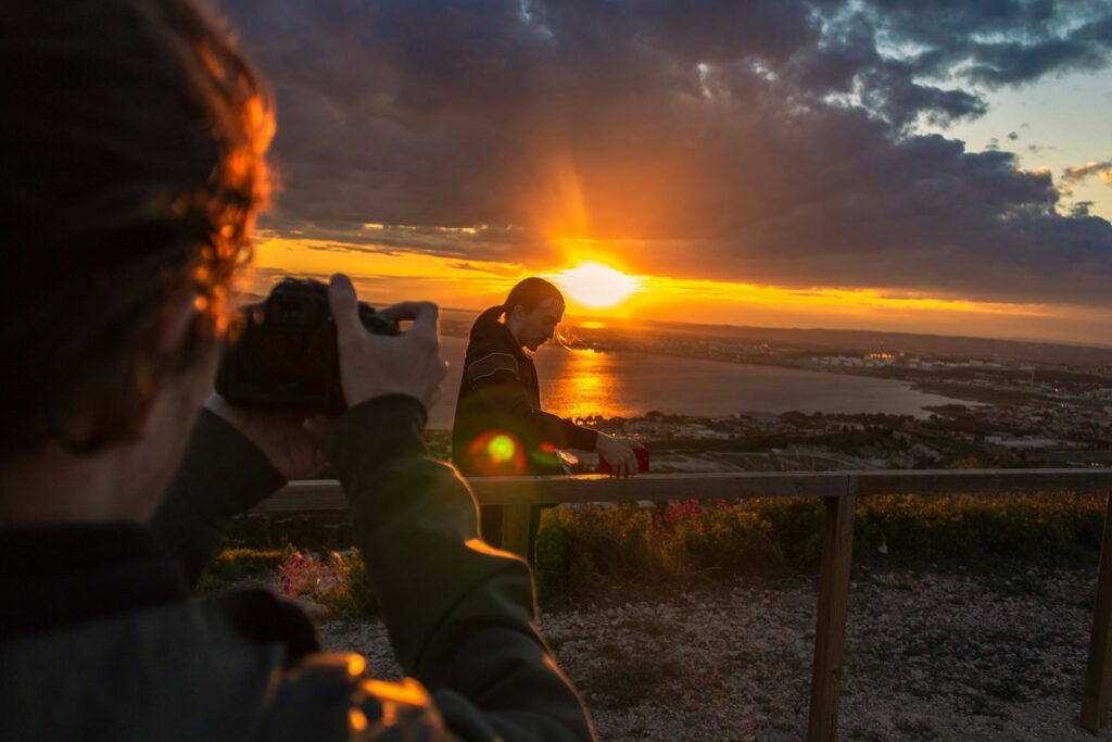 A person is using a DSLR camera to take a photo of another person at sunset. 
