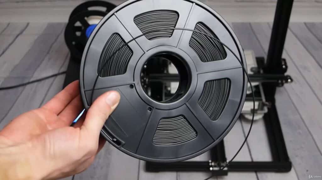 A person’s hand holding a large spool of black plastic filament in front of a 3D printer sitting on a gray striped tabletop. 