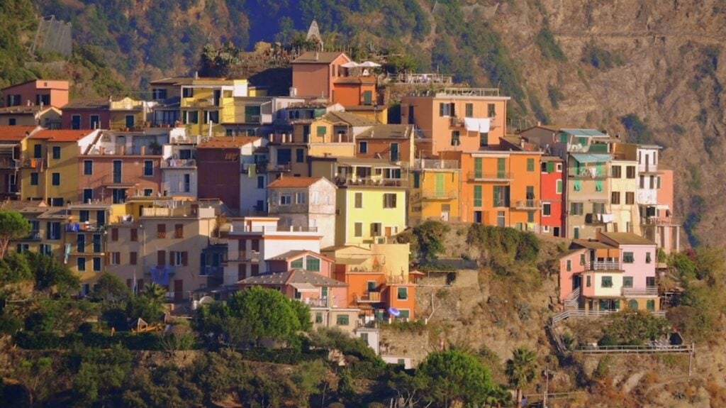 A group of colorful multi-story houses built on a hillside dotted with green shrubbery. The houses are painted various shades of orange, yellow, red, pink and green. 