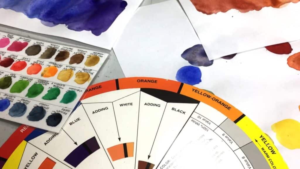 A round color chart and several sheets of white paper covered with multicolored watercolor swatches, all strewn on a gray tabletop. 