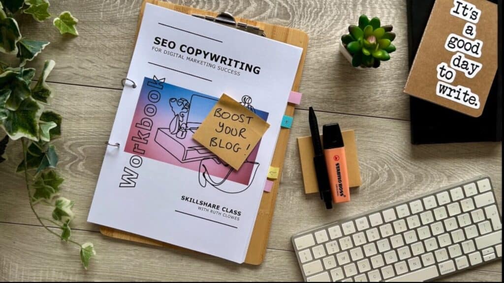 A workbook titled 'SEO copywriting for digital marketing success' laying on top of a wooden clipboard on a wooden desktop. The front of the workbook is affixed with a sticky note which reads 'boost your blog!', and other visible items on the desk include a keyboard, plants, pens and notebooks.