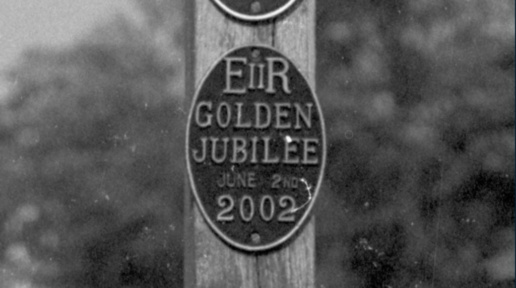 Two black and white vintage photographs of an oval, metal sign on a wooden post sit side by side. The photo has multiple dust spots and scratches that obscure the lettering.