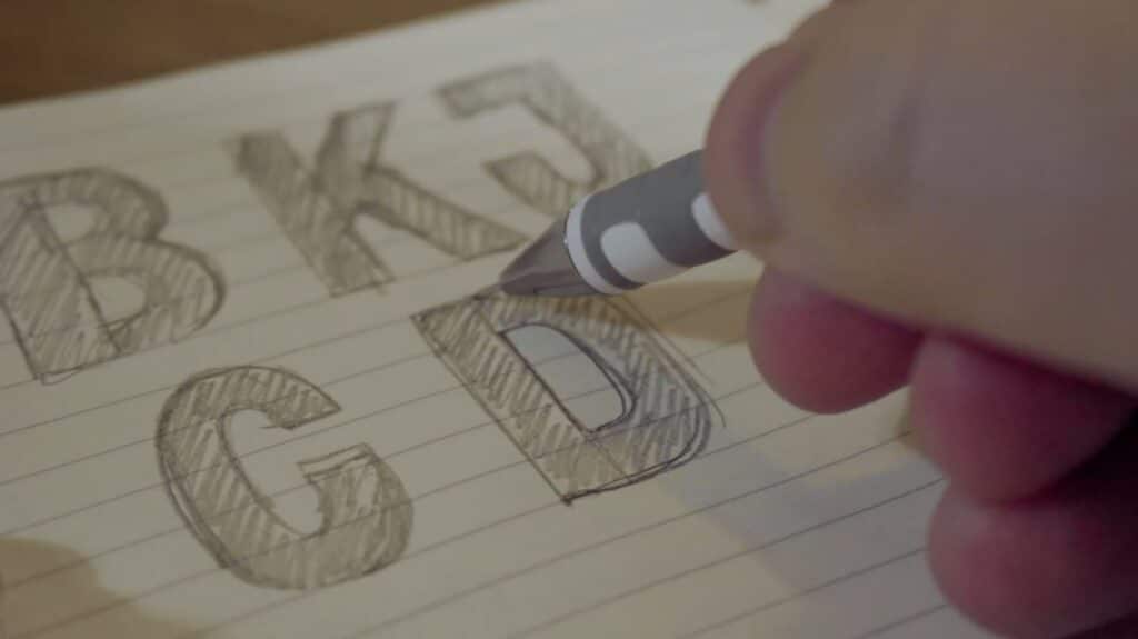 A close-up shot of a person’s hand using a mechanical pencil to sketch the capital letter ‘D’ on a piece of lined paper. Other hand-sketched letters are also visible on other parts of the paper. 