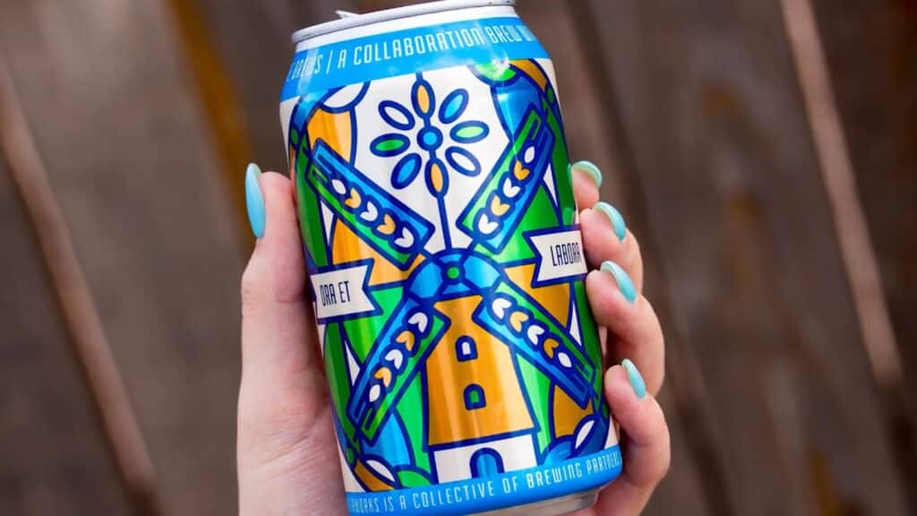A woman’s hand holding a beer can in front of a brown wooden background. The beer can is decorated with a blue, orange, green and white design that resembles a windmill, and the woman’s fingernails are painted a bright shade of turquoise. 