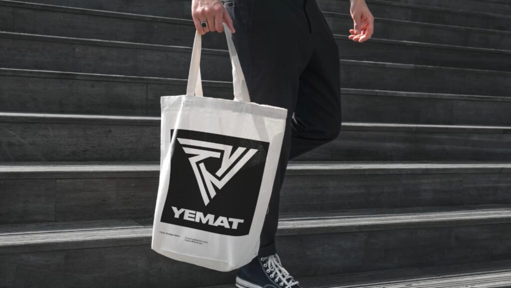 A man in black pants and Converse sneakers walking down gray concrete steps. He’s holding a white canvas tote back that’s decorated with a large black and white logo that reads ‘Yemat.’