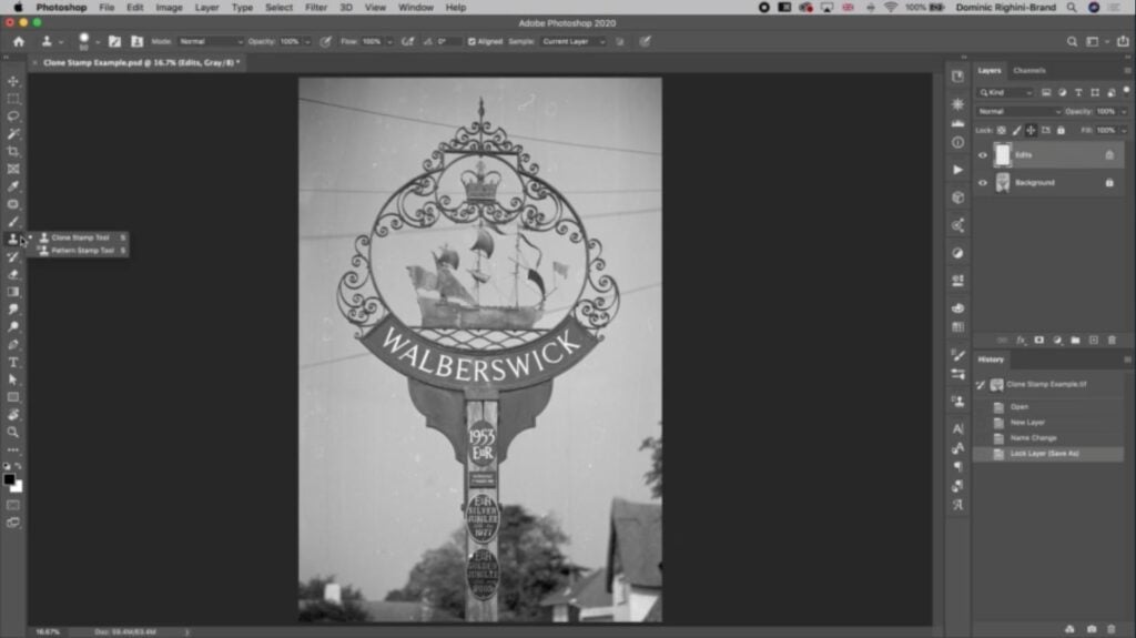 A black and white vintage photo of an old iron sign that reads “Walberswick” is open in Photoshop. The cursor is hovering over the toolbar to the left of the screen and a menu is open with two options: Clone Stamp Tool and Pattern Stamp Tool. 