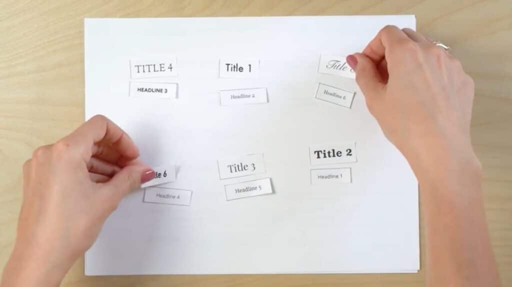 A white piece of paper with 12 small rectangular pieces of paper laid on top of it. Six are labeled ‘Title 1’ through ‘Title 6’ and are printed in various fonts, and six are labeled ‘Headline 1’ through ‘Headline 6’ and are printed in various fonts. A person’s hands are in the process of picking up two of the ‘Title’ pieces to swap them.