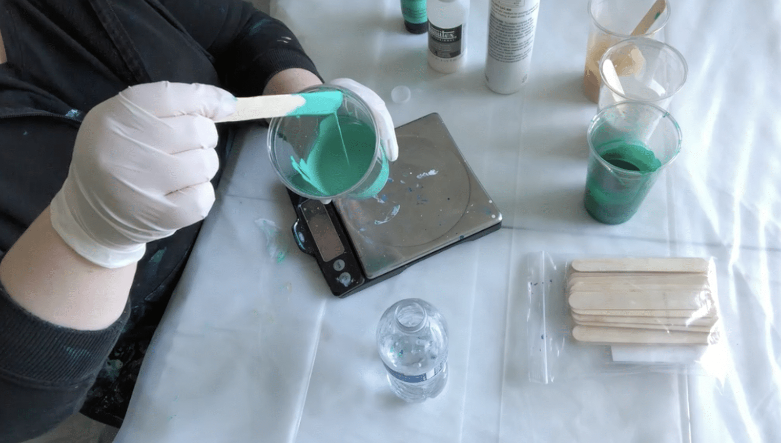 Lady with glvoes mixing cup of teal paint. On the table below is a few other cups of paint, water, and a scale.