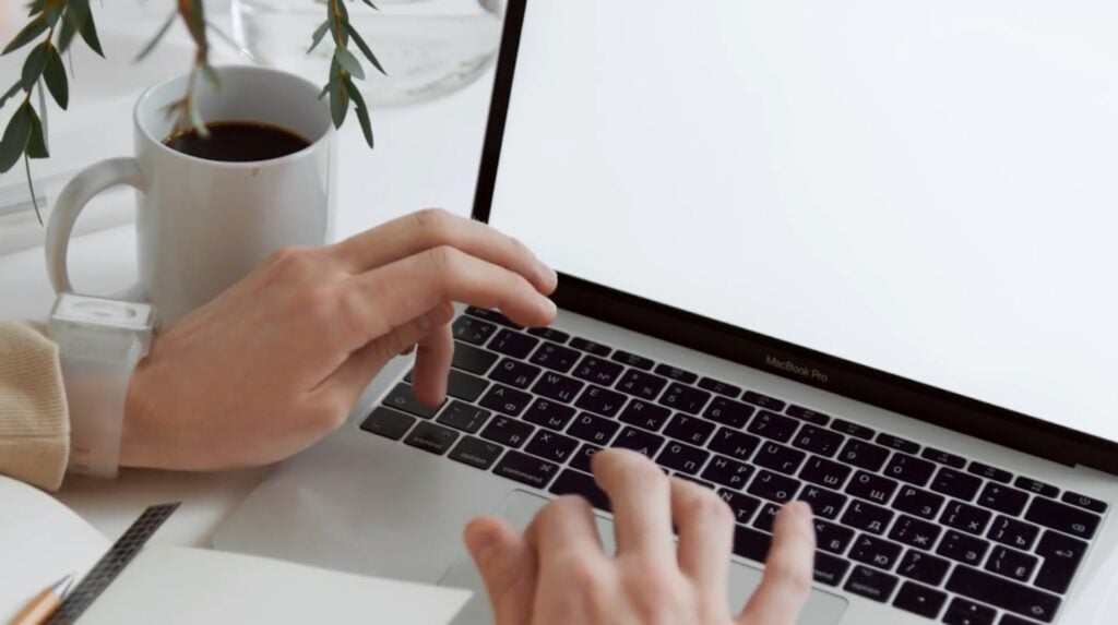 A person’s hands hovering over the keyboard of a Macbook Pro laptop resting on a white table. To the left of the laptop is a white mug full of coffee, along with the green leaves of a nearby plant.
