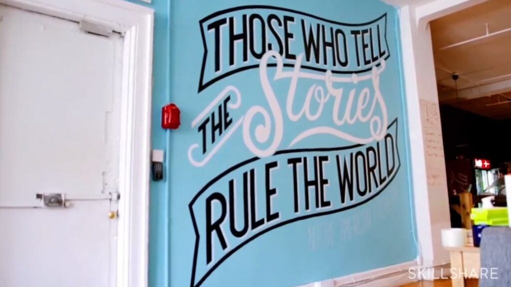 A blue wall next to a white door. The wall is painted with a mural that reads ‘those who tell the stories rule the world’ in black and white text.