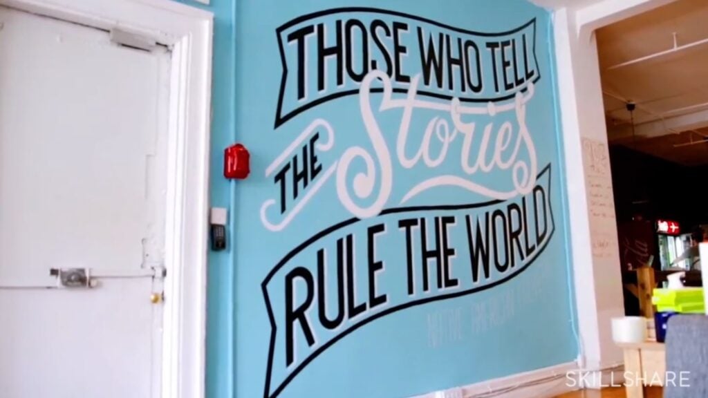 A blue wall next to a white door. The wall is painted with a mural that reads ‘those who tell the stories rule the world’ in black and white text. 