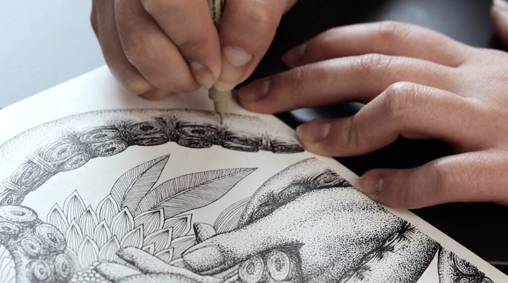 An artist adds blank ink dots to a stippling drawing of an octopus’ tentacle. The entire image isn’t visible but a hand and some greenery can also be seen around the tentacle.