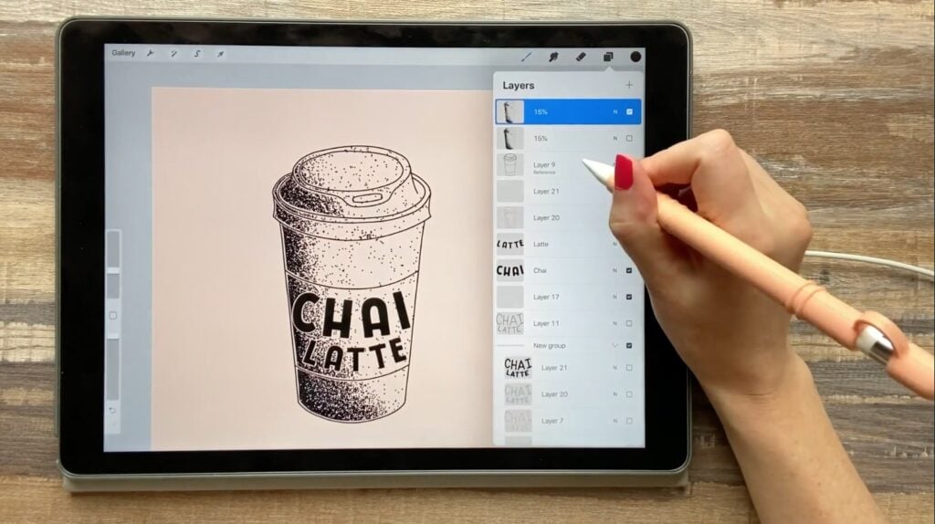 An iPad open to Procreate sits on a wooden surface. Procreate is open to a beige background containing a drawing of a chai latte in a to-go cup. The artist has the layers open and is hovering their stylus over one of the layers.