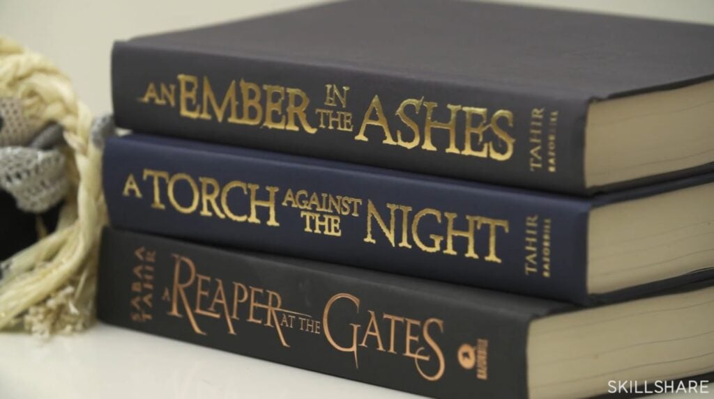 A stack of three hardcover books sitting on a white tabletop. The top book is titled ‘An Ember in the Ashes,’ the middle book is titled ‘A Torch Against the Night’ and the bottom book is titled ‘A Reaper at the Gates.’