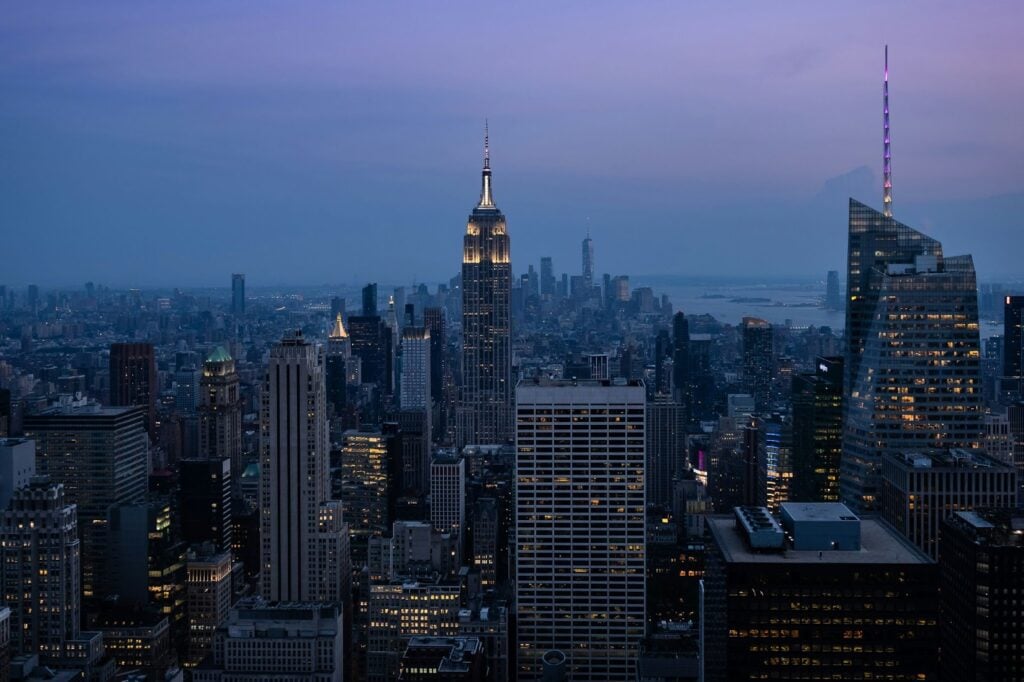 New York City skyscrapers cover the horizon during blue hour. The sky is a muted blue and purple and lit-up windows cover the buildings.