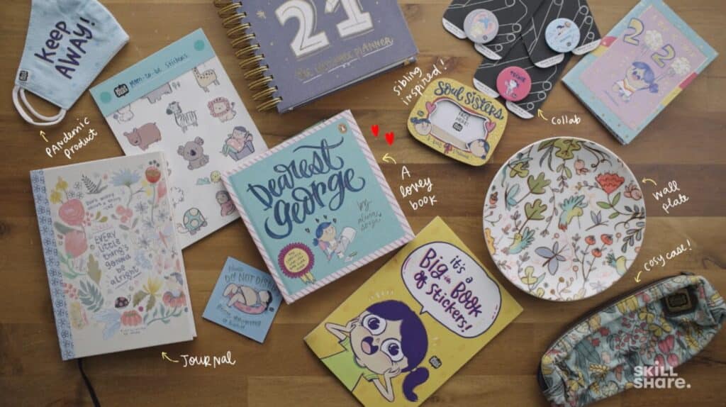 Various pieces of merchandise strewn across a wooden tabletop, including a plate, greeting card, journal, face mask, pencil case and stickers.