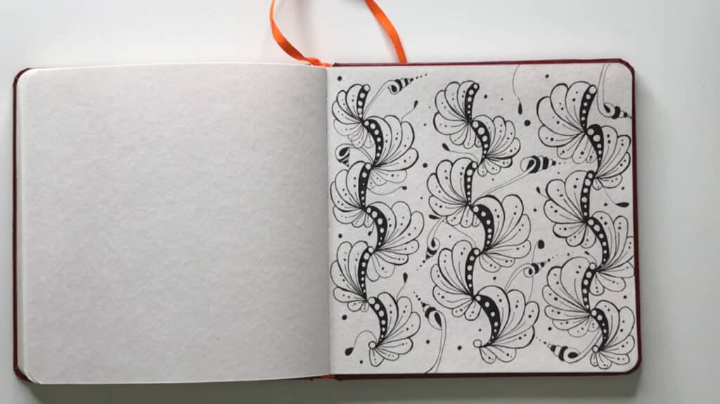 A sketchbook laying open on a white tabletop. Its page is filled with ink doodles that resemble scalloped pasta. 
