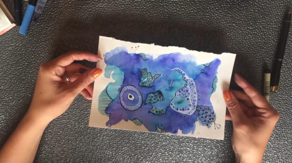 A person’s hands holding a white piece of paper filled with blue and purple watercolor paint that’s accented with ink doodles. 
