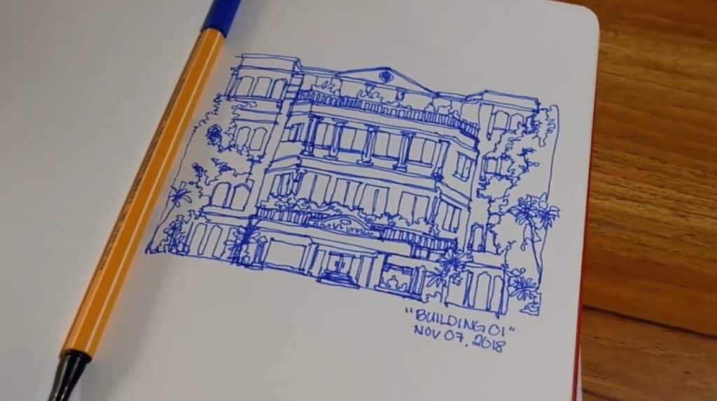 Alt: A sketchbook lying open on a wooden tabletop. One page is filled with a blue ink drawing of a multi-story building. 