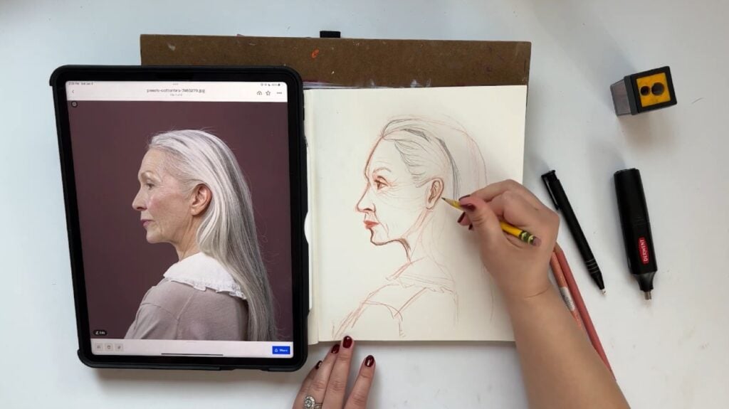 A person using a pencil to draw the side profile of a woman with long gray hair on white paper. An iPad displaying a reference photo is visible to the left.